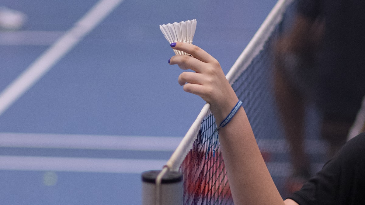 A player's hand holding a shuttlecock over the net on an indoor badminton court, with the boundary lines visible on the court