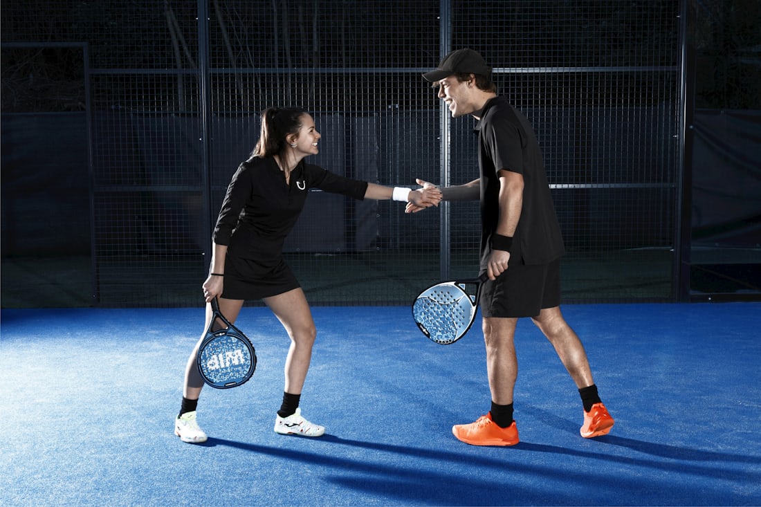 Two players with padel rackets greeting each other inside a glass-walled, mesh-fenced padel court — the social doubles game new players start with.