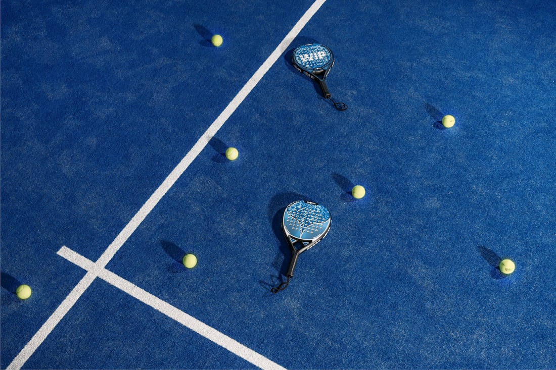 Two padel rackets and several balls scattered across a blue padel court — the gear you go through fastest as you start playing weekly.