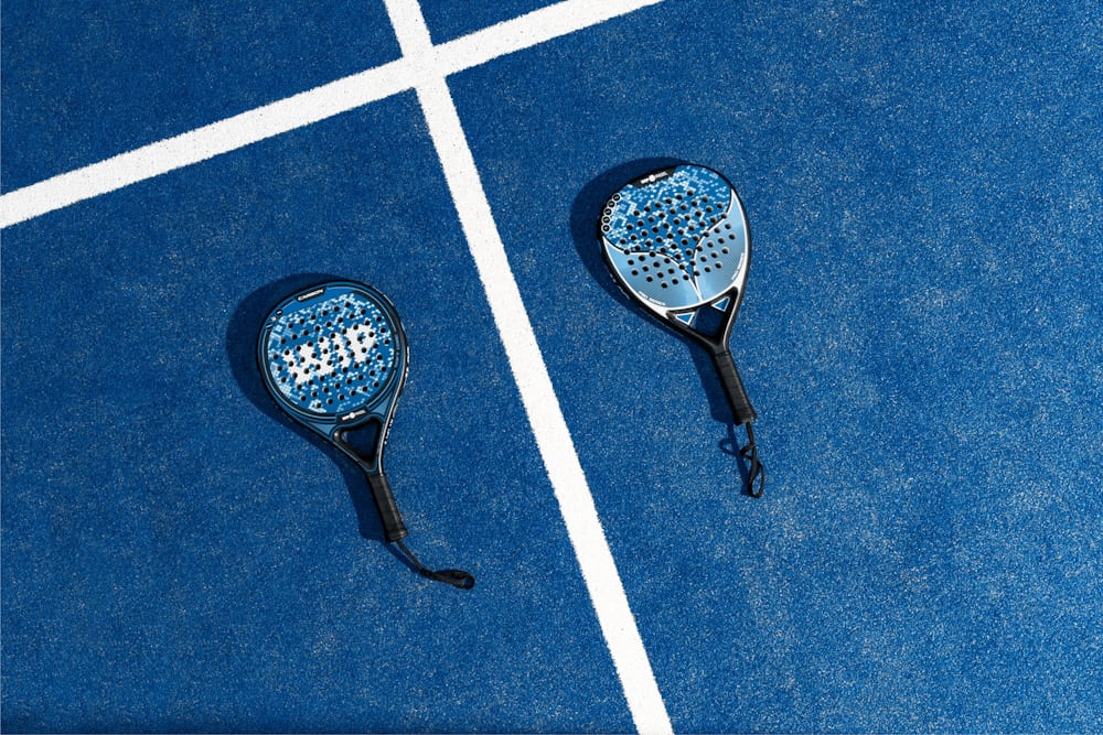 Two padel rackets laid out on a blue court — the solid, perforated paddle faces that define the sport.