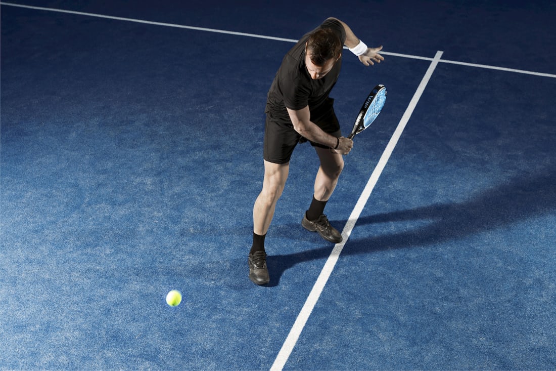 Jugador a media volea con una pala de pádel en una pista azul, con el juego de pies y las zapatillas en cuadro: el movimiento que exige un calzado diseñado específicamente para el pádel.