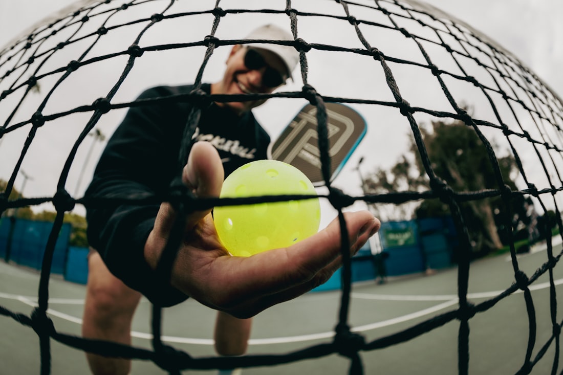 A pickleball player holds a yellow outdoor pickleball with the net and court visible behind them