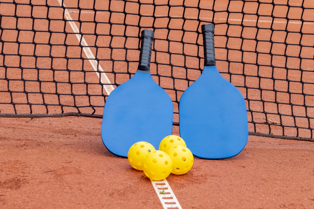 Two pickleball paddles and three balls resting on the sideline of an outdoor pickleball court