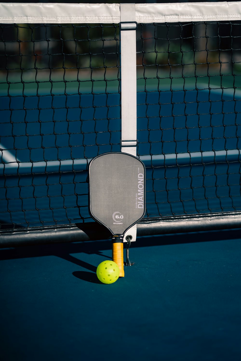 Court-level view of a pickleball ball next to a paddle on a freshly painted blue and green court