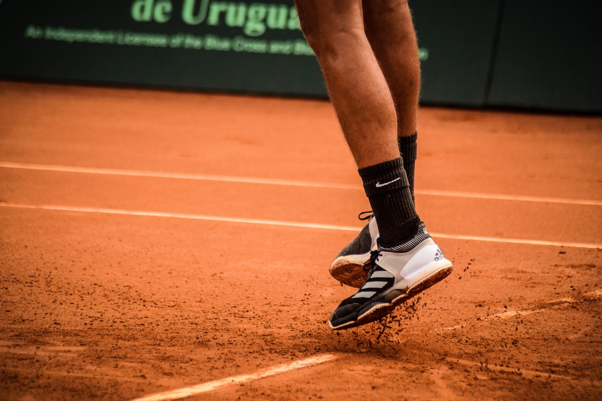 A tennis player's feet, mid-jump in white and black tennis shoes, kicking up clay-court dust near the baseline