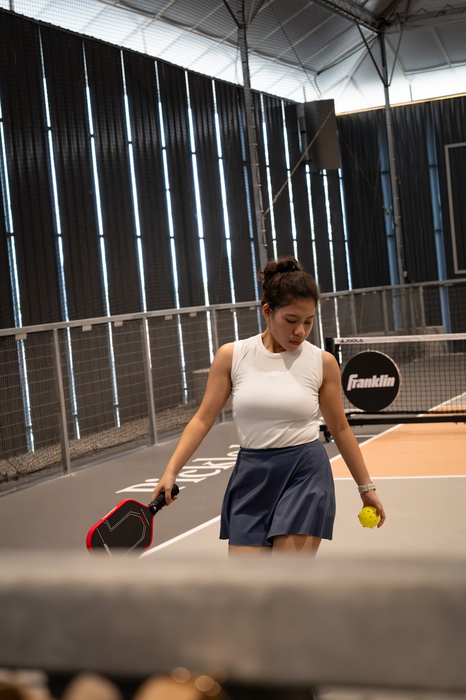 Una jugadora sostiene una pala y una pelota en una pista de pickleball cubierta, con las paredes de lona y los tragaluces del recinto visibles detrás