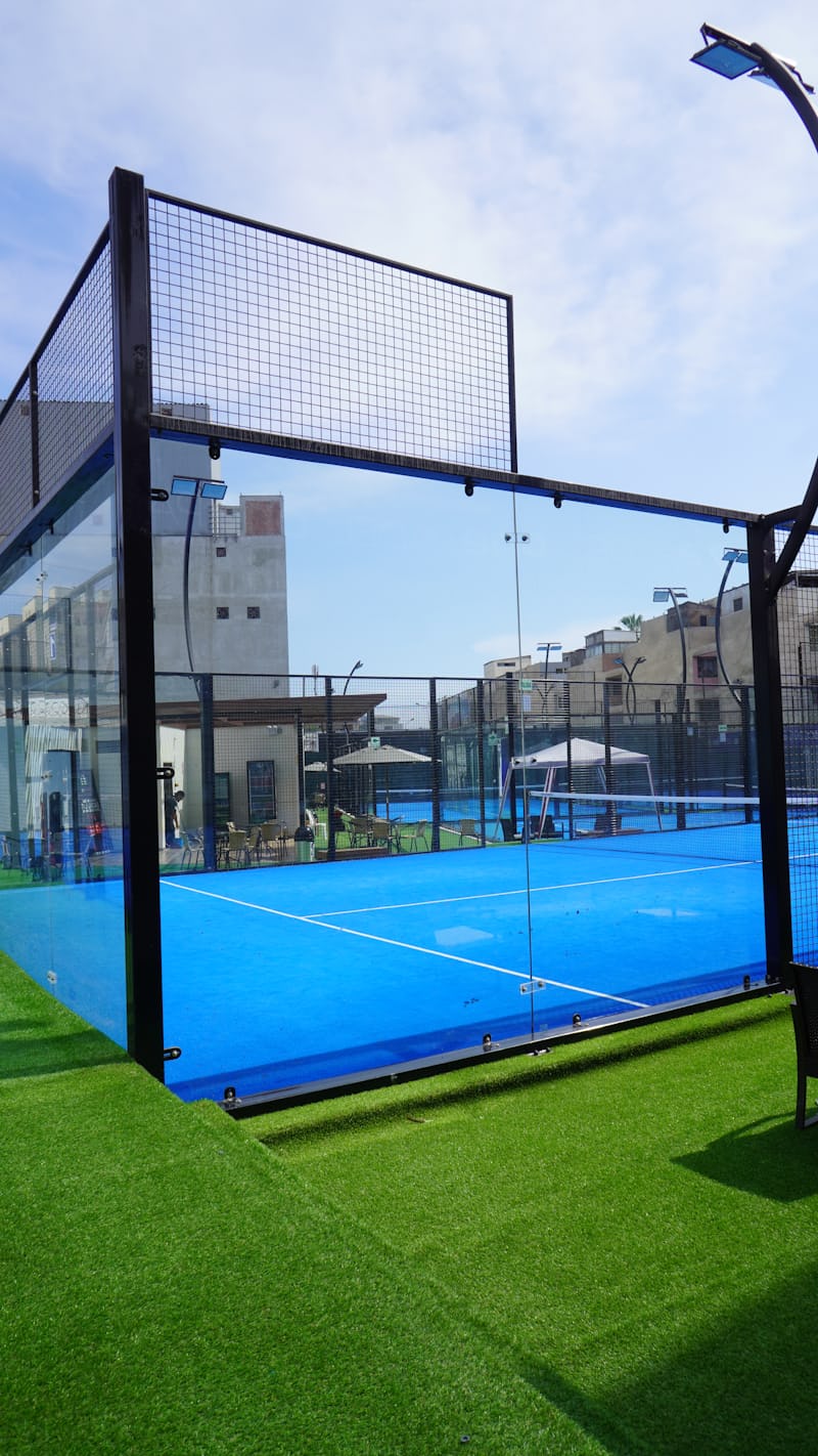 Una pista de pádel con paredes de cristal y malla metálica y una superficie de juego azul brillante, vista desde detrás del cristal con un club social y otras pistas visibles al fondo
