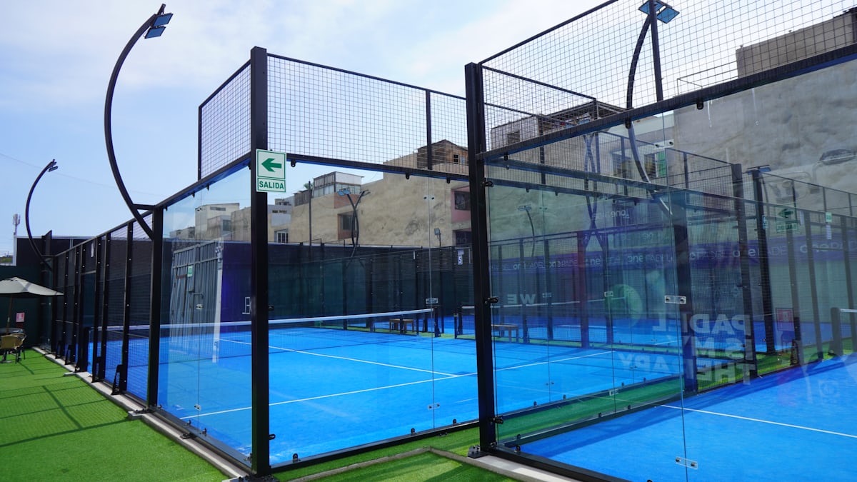A padel court corner showing the glass walls topped with metal mesh fencing that define the sport, with the blue court surface visible beyond.
