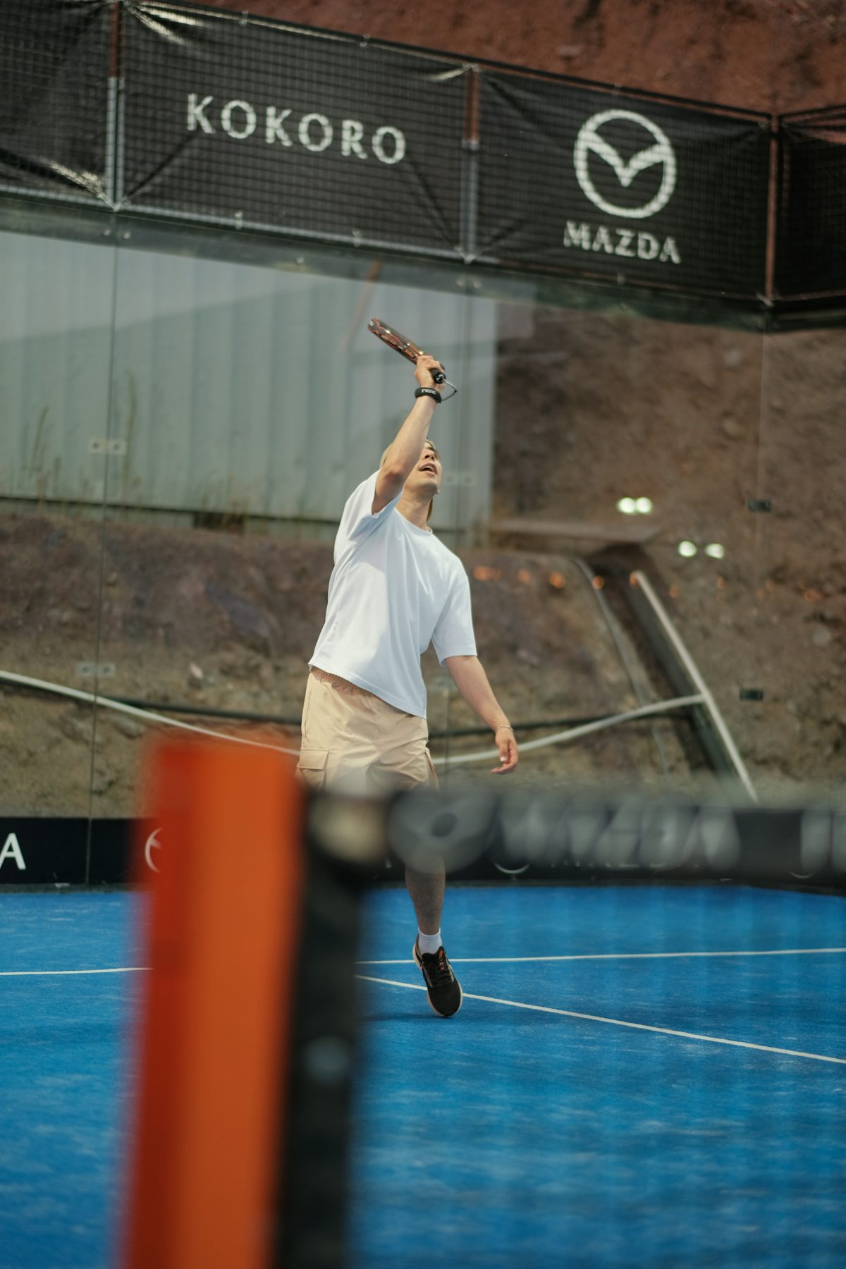 Un jugador de pádel se estira hacia arriba con la pala levantada por encima de la cabeza para golpear un remate, con la pierna delantera elevada en pleno movimiento sobre una pista azul