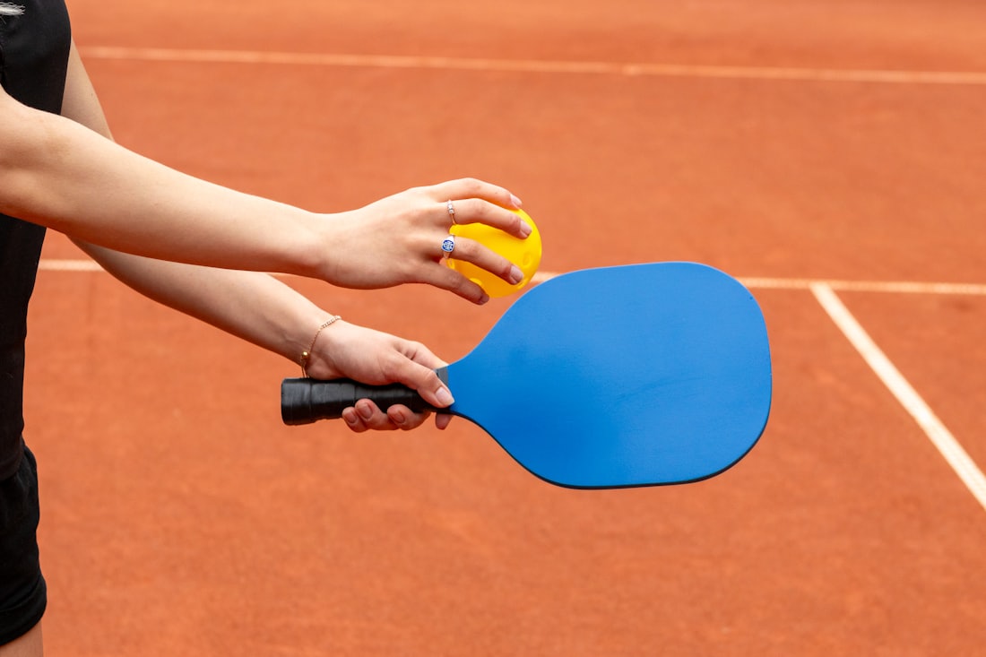 Una persona sujeta una pala de pickleball y una pelota en la línea de fondo de una pista exterior soleada