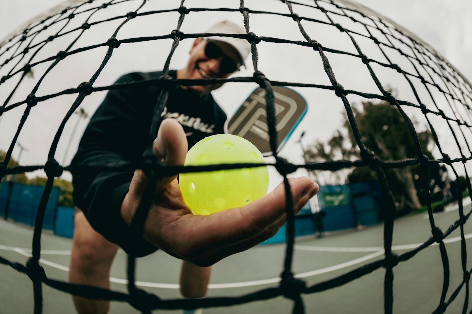 A pickleball player smiles while holding a bright yellow pickleball out toward the camera through the net, with a paddle visible over their shoulder on an outdoor court