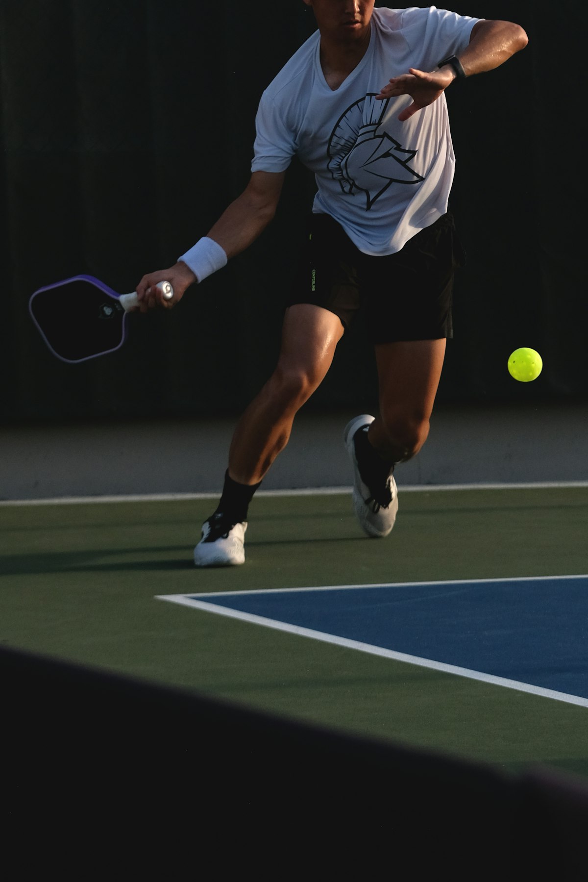 A pickleball player mid-swing on court, paddle extended toward an airborne yellow ball near the non-volley zone line