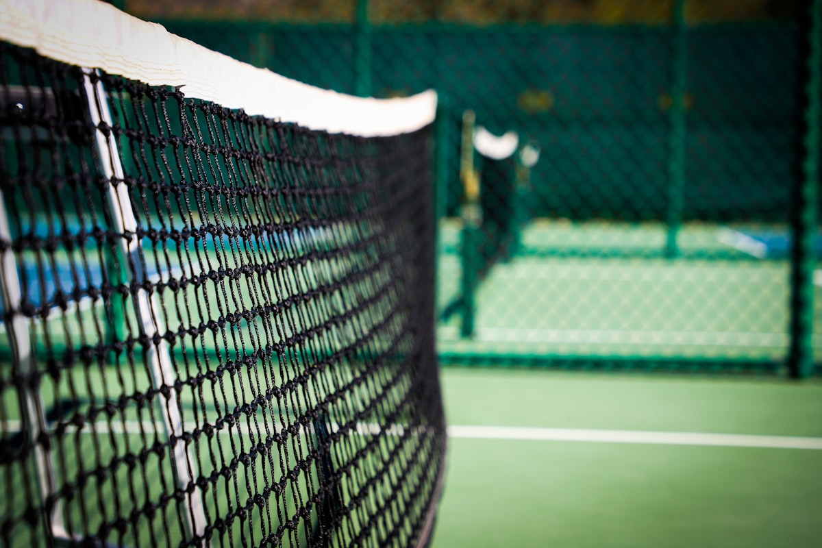 Close-up of a pickleball net's top band and mesh, with the court and far net post visible in soft focus behind it