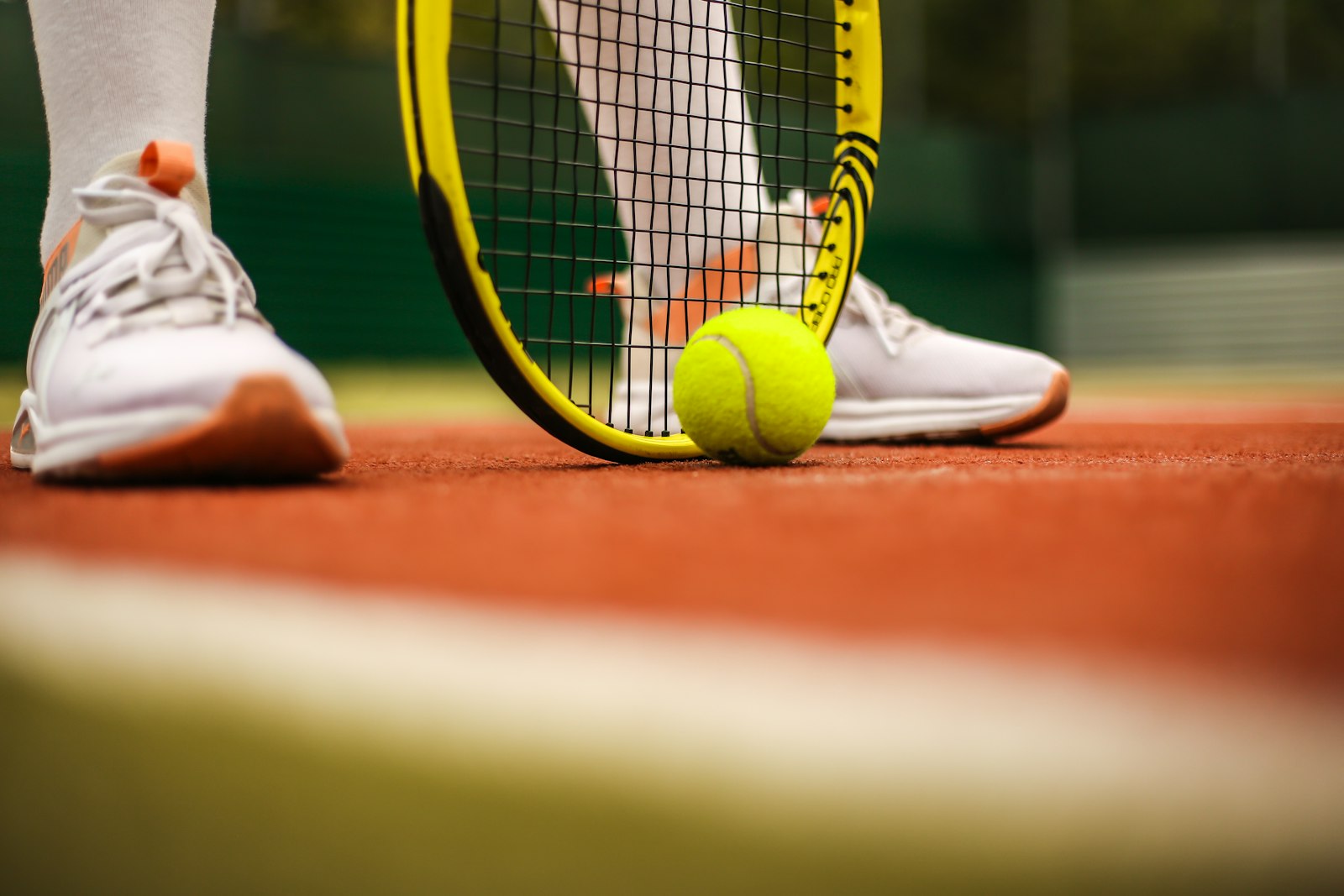 Close-up of a tennis player's white sneakers, racket, and ball at court level on a clay court, illustrating footwork and court positioning