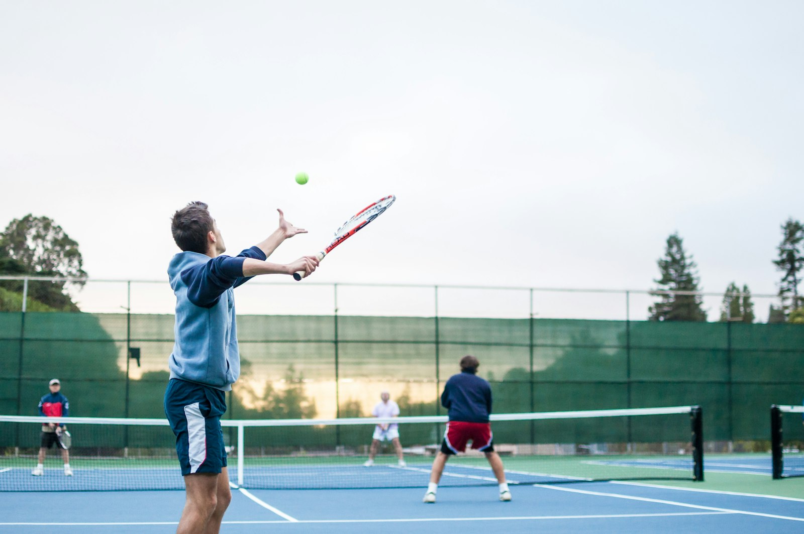 Four players on a tennis court during a doubles match, one serving while his partner and both opponents hold net position