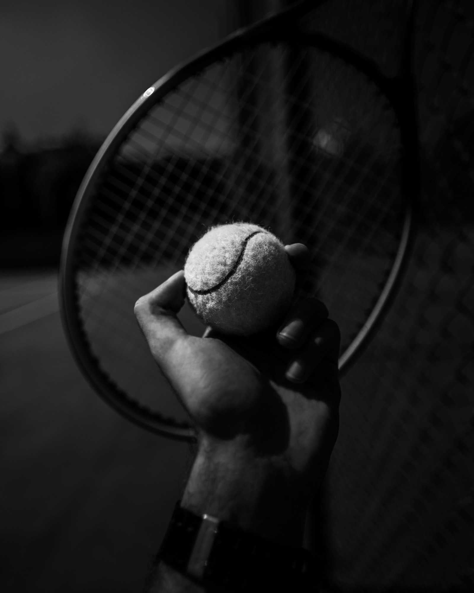 Black-and-white close-up of a hand gripping a tennis ball in front of a racquet's strings